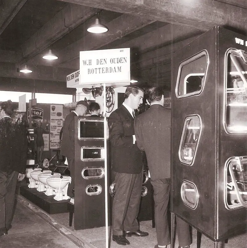 Visitors in suits examine a row of marine toilets and portlights at the same 1960s W.H. Den Ouden booth, the company's Rotterdam signage mounted overhead on a pole.
