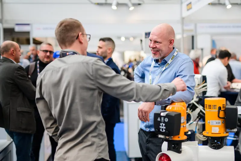 Two men shake hands at a trade show stand, black and yellow bow thruster motor housings stacked on the table between them.