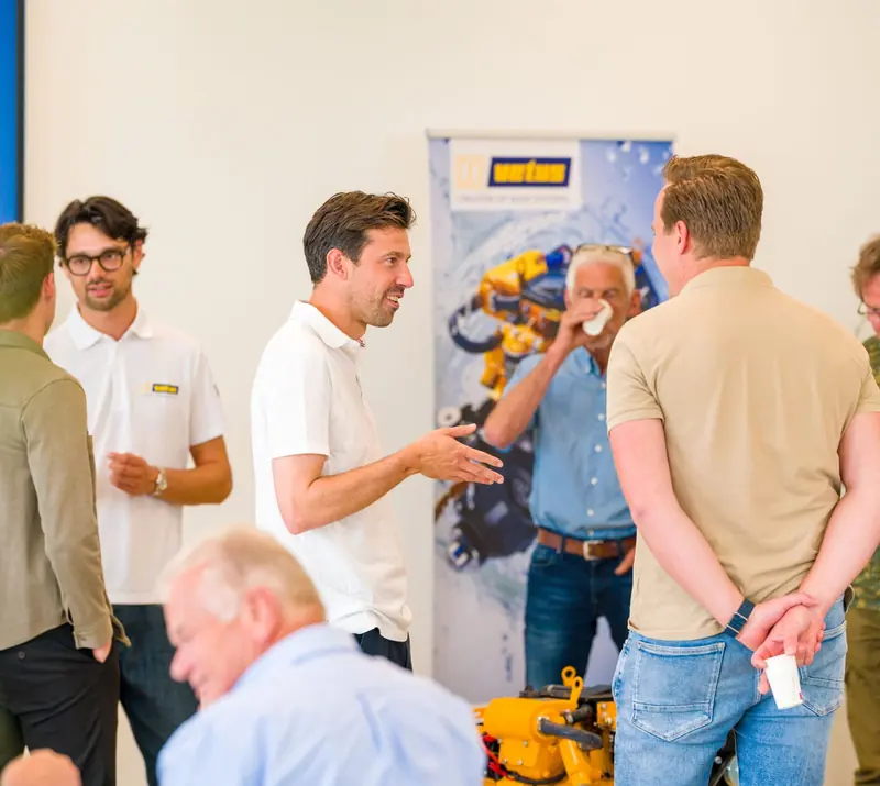 A small group talks beside a VETUS pull-up banner showing a yellow thruster unit, another real yellow thruster sitting on a table in the foreground.
