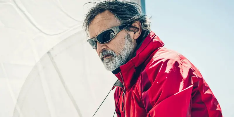 A grey bearded sailor in an offshore jacket looks out from the rail as the headsail fills behind him.