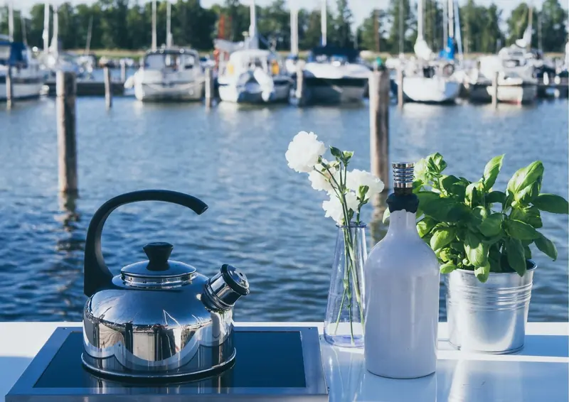 A stove top kettle sits on a boat's built in hob at a marina berth, basil and cut flowers alongside.
