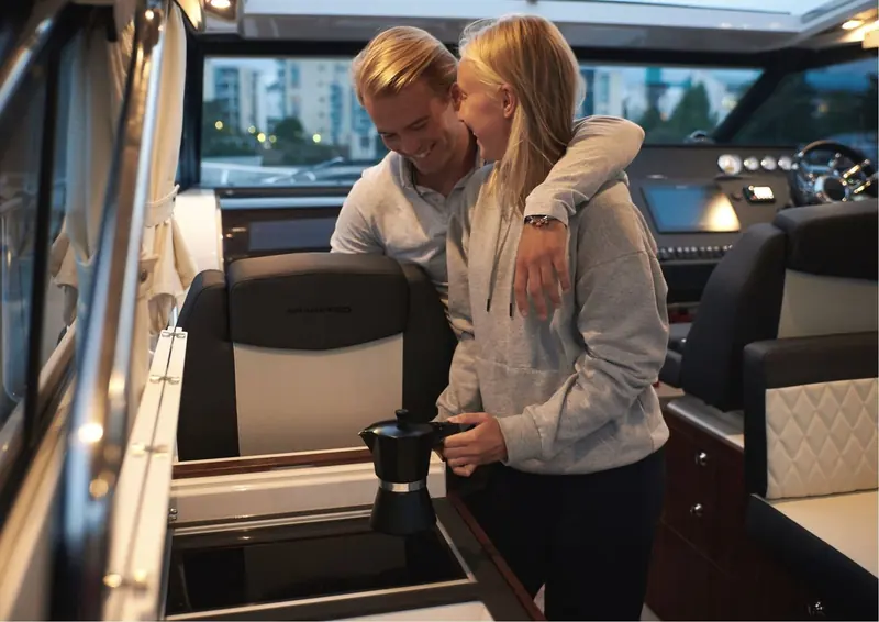 A couple make coffee on a stovetop moka pot at the galley hob of a Grandezza motorboat, the helm visible behind them.