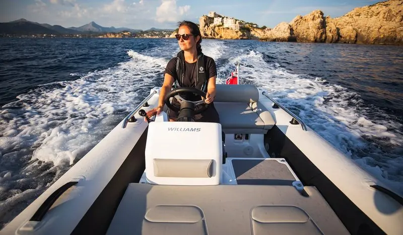 At the wheel, a driver runs a Williams tender out past a rocky headland with the wake curling behind.