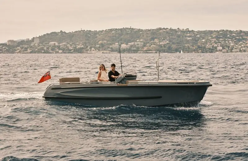 A couple cruise slowly in a grey open tender off a wooded coastline, red ensign flying aft.