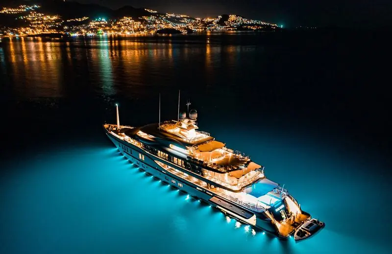 Underwater lights turn the sea turquoise around a large motor yacht anchored off a lit coastline.
