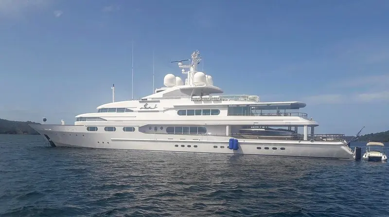White and still at anchor, a large yacht sits off a wooded shore with her tender astern.