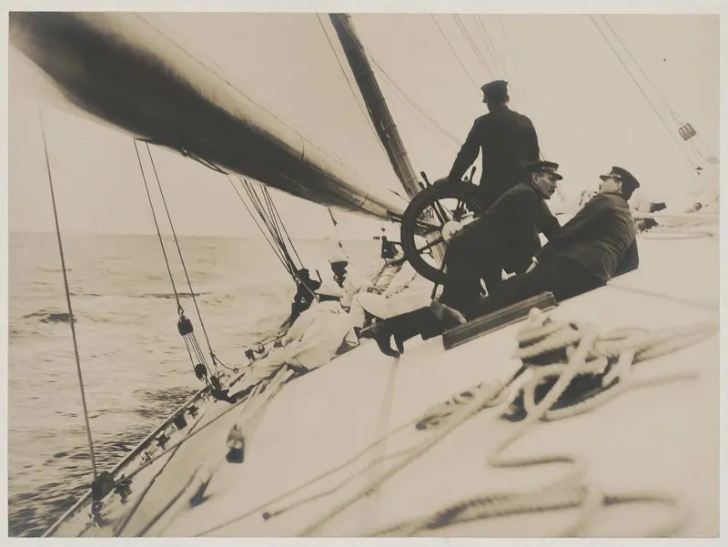 The afterguard of the racing yacht Reliance work the wheel on a heeled deck, 1903