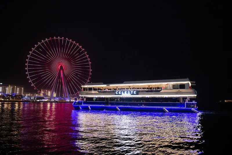 An illuminated dinner-cruise boat passing the Ain Dubai observation wheel at Bluewaters Island by night