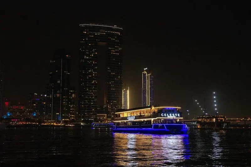 A lit dinner-cruise boat off Dubai Harbour at night, residential towers glowing in the background