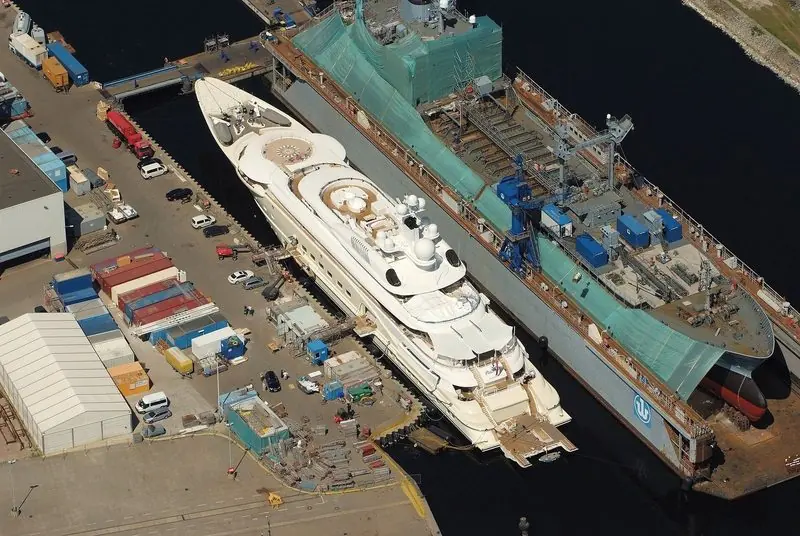 Seen from the air, a large motor yacht lies alongside at Wilhelmshaven with a naval hull in the floating dock beyond.