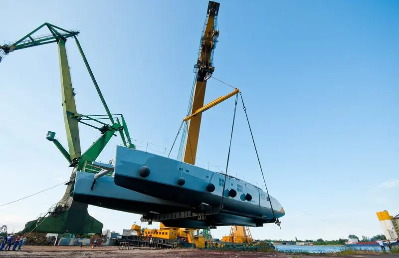 A catamaran hull hangs in the slings between shipyard cranes at Gdansk.