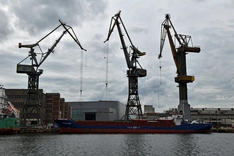 Three of the shipyard's cargo cranes lift steel sections into a hull under a grey sky.